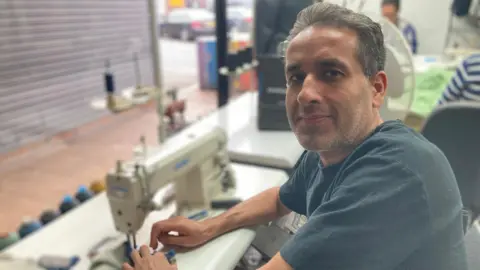 BBC Sabur Kirimi wearing a blue tshirt and looking at the camera is sat in front of a sewing machine