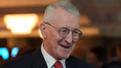 PA Media Hilary Benn smiling. He wears a black suit, white shirt and red tie. He has grey hair.