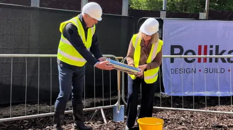 Two people hold a metal time capsule tube. Both wear high vis yellow vests and hard hats. They are leaning in to place it down into the ground. They stand in front of metal hoardings for building sites.