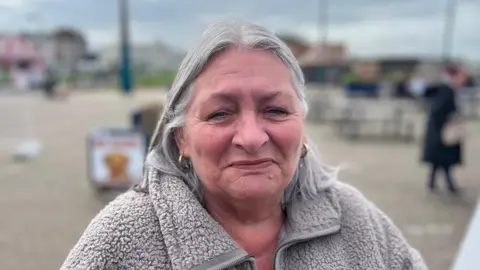 Linda Williams, who has grey mid-length hair, smiles for the camera. She is wearing a grey fleecy jacket.