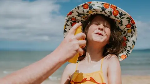 A little girl on the beach wearing a sun hat and orange swimming costume grimaces as sunscreen is applied to her face. 