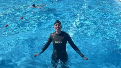 CamSight Lucy is standing in the lido withe the water up to her waist and her arms stretched out to her sides. She is smiling at the camera and the water is a bright blue. She is wearing a black wetsuit - with Zones written in the centre - and a black swim cap with goggles on her head.