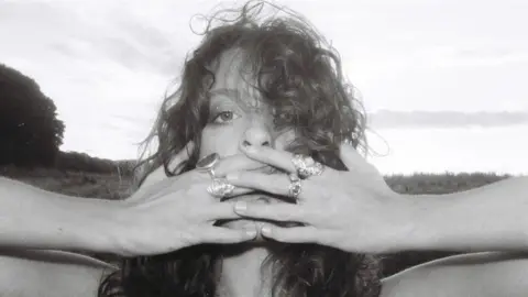 Heid Curtis has curly hair and is covering her mouth with both hands and is wearing a number of rings. The photo is black and white and there is a field and trees behind her