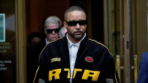 Stylist Deonte Nash, wearing a black and yellow jacket, blue shirt underneath and sunglasses, exits the courthouse. 