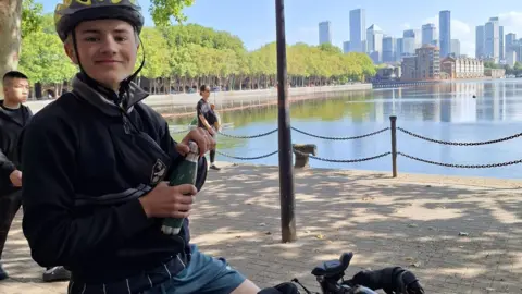 Victoria Bacon Ranulph, wearing a full-sleeve navy shirt, navy shorts and a yellow helmet, sitting on a cycle. He is holding a green water bottle while smiling for the camera.