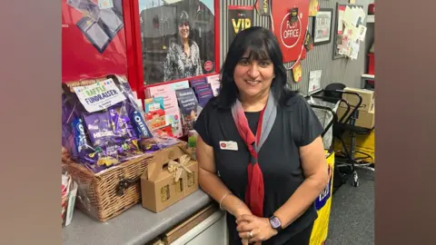 Roopie Gill standing behind a Post Office counter beside a raffle fundraiser display featuring a wicker hamper, chocolates, biscuits, and gift items. A large poster in the background and displays a photo of a staff member. The shop interior includes Post Office signage, product brochures, and community noticeboards.