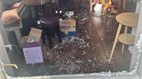 Finn Reeves The photo peers through the front door of a pub where the glass has been broken in. There is glass all over a brown wooden floor. There's a box of Corona Extra beer in the centre of the image, with pub stools on both edges of the frame.