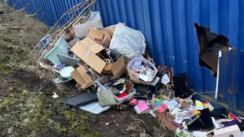 Anna Woodman Lots of household and personal belongings strewn across the green bank at the back of the blue tin unit. Amongst the stuff, we can see a PC screen, a handbag, brown boxes, some books and a photo frame. Can also see a big cut out square peering into the tin.