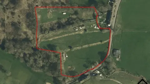 LDRS Aerial photograph of a semi-rural property outlined in red. The plot includes grassy fields, scattered trees and several buildings. A road runs along the right edge, with additional structures nearby. Wooded areas border the left side.