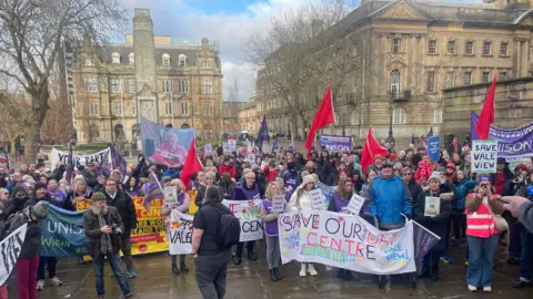 Some hundreds of people gather with various banners and flags calling for care venues to be saved. They stand in a large square in Preston city centre in front of historic grand buildings.