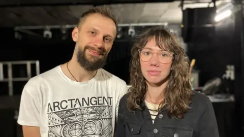 Adam and Sarah from Life in Mono - he is wearing a white t-shirt that says Arctangent on it in black with a black design underneath. She is wearing a black boiler suit and has glasses