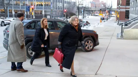 A woman wearing a long black coat with a red bag in hand is walking into a courtroom as a male and female follow her in from behind. 