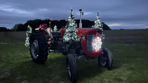 A red tractor in a field in slightly dark conditions, it is lit with fairy lights and decorated with tinsel 