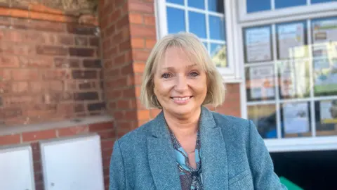 BBC News A blonde lady with a bob and a fringe, she's smiling at the camera with gold hoop ear rings in her ears and a blue blazer stood behind the brick walls at the Cherry Tree Centre in Beverley