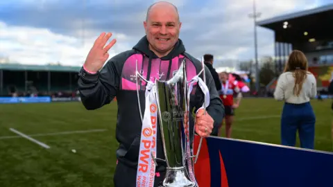 Sean Lynn holds the Premiership Women's Rugby trophy and three fingers up to symbolise their three consecutive titles