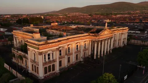 A still image taken from aerial drone footage of the former Crumlin Road Courthouse in north Belfast.  The large Victorial building has a Palladian portico but most of its roof is missing and there are scorch marks from previous arson attacks. 