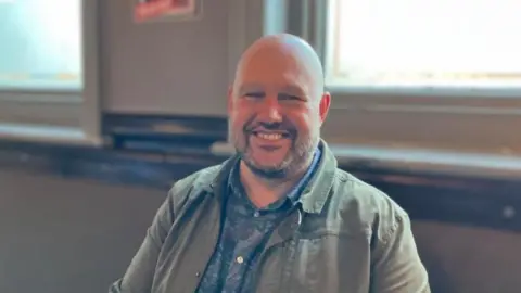 A bald man with a short beard wearing a khaki jacket