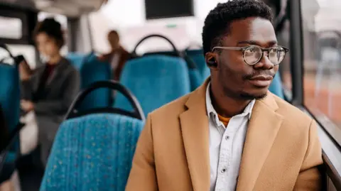 Man wearing glasses, sitting by the window on a bus, looking out the window while listening to music with wireless headphones. He's wearing a while shirt, with a smart cream coat.