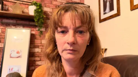 A lady with ginger hair and a fringe, she has black glasses on her head and is sat in a living room. The lady has pale skin and blue eyes and is wearing an orange jumper. 