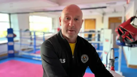 Un hombre de cabeza calva, con un top negro, mira la cámara. Detrás de él hay un anillo de boxeo