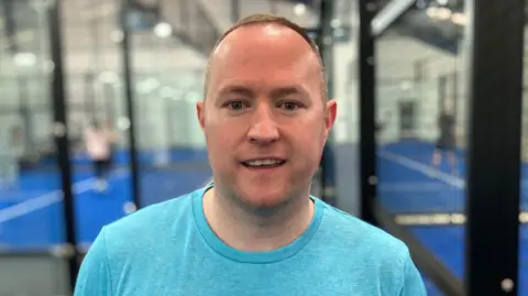 Sam Read/BBC A man in a light blue shirt smiles at the camera while padel is played behind him.