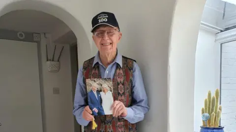 Family photo Peter Byers is wearing a black cap with the number '100' on it. He is wearing a multi-coloured tapestry waistcoat over a blue and white striped shirt. He is holding a card with a photograph of King Charles and Queen Camilla on it.