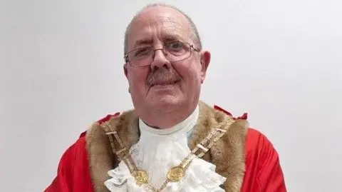 Newcastle-under-Lyme Borough Council A bald man with glasses and a moustache smiles while wearing red robes and a mayoral chain. He is in front of a white wall.