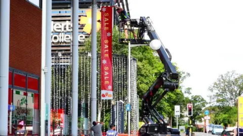 Stephen Danzig A man on a cherry picker is fixing a sign advertising a sale to the outside of West Herts College near some Christmas lights