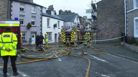 BBC Firefighters at the scene of the fire in Kirkby Lonsdale. Several firefighters are working around an engine. Several houses have been extended from the back of the engine. The road has been cordoned off and a police man stands to the left.