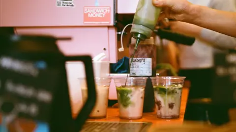 Alex Douglas Iced coffees sit in the background on a counter in an orange store.