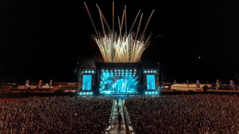 At the centre of the photo is the main stage lit up in blue with fireworks being fired off behind. In front of the stage is a sea of people 