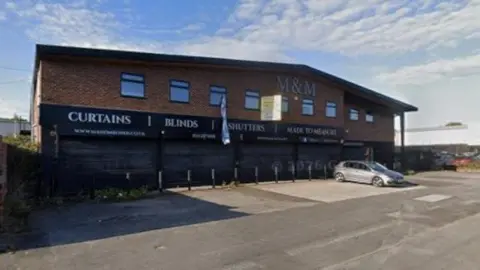 A two‑storey commercial building made of red brick, positioned along a wide paved road. The building has a slightly sloped roof and several rectangular windows evenly spaced along the upper floor. Across the ground floor, large black shuttered fronts display prominent white lettering that reads “CURTAINS”, “BLINDS”, “SHUTTERS”, and “MADE TO MEASURE”.