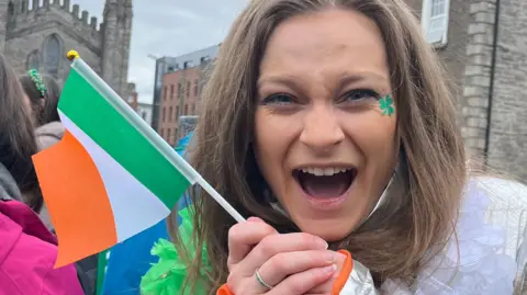 Sam Kwan Emmy smiling and holding a Republic of Ireland flag. She has a shamrock sticker close to her left eye