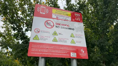 Red and white warning sign with trees and bushes behind. The sign says Dim Nofio/No swimming and has a forbidden to swim sign of a swimmer with a red line over to warn people. It also warns of other risks including fast currents and hidden or submerged objects
