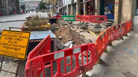Simon Thake/BBC A whole in the ground on a city centre street with mess and red fences around it