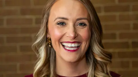 A woman with long blonde hair and gold earrings smiles at the camera