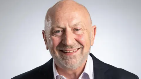 Blue Light Whistle Blowers A close‑up portrait of an older, bald man with a short grey beard, wearing a dark suit jacket over a light checked shirt, set against a plain light grey background.