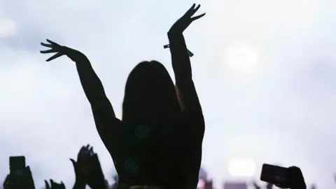 A general view of the silhouettes of people at a concert. Some have their phones up and arms in their air, with one person on someone's shoulders with their arms outstretched. Lighting and stage smoke can be seen in the distance.