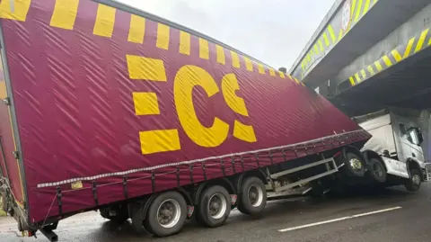 Hinckley Town Centre (Facebook) Image shows a red lorry with a white front and yellow writing on the red main section. The front park of the load is wonky as that is the section that has hit the bridge, and the vehicle is now stuck under the bridge, with the driver section lifted from the road surface. 