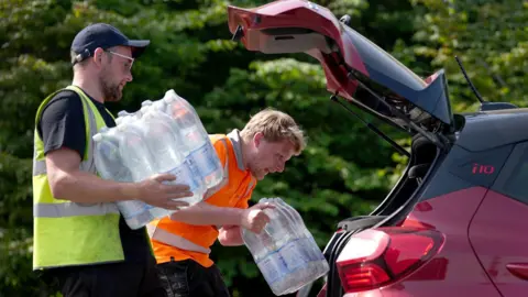 A man wearing a yellow his vis jacket and a man wearing an orange hi-vis jacket, each carrying a pallet of water, are in the process of putting it into the back of a red car.