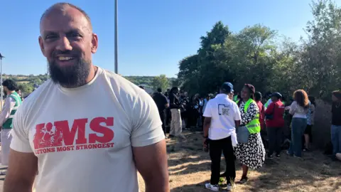 Louise Parry/BBC Tariq smiles at the camera, standing in the park with crowds gathered behind him. He wears a white T-shirt saying "LMS Lutons Most Strongest".