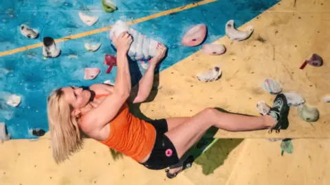 A woman with blonde hair is on an indoor climbing wall, surrounded by assorted-shaped and assorted-coloured plastic lumps attached to the wall. She is holding herself against the wall, with both of her hands holding a larger blue, ridged lump, with pink spots. She is wearing an orange top and black shorts. Her shoulder-length hair is hanging down with gravity.