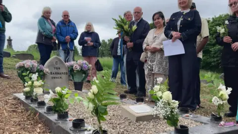 A picture of the grave, the gravestone has the words C754 Barnardo Owner engraved on it, there are 15 plaques with the names of each child, there are cream flowers and people gatherered around it.