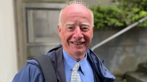 Deputy George Oswald, a man with a bald head, but white hair on the sides of his head. He's wearing a blue rain coat, over a blue shirt and a blue and white tie.