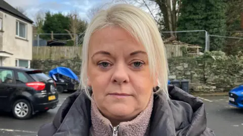 Charlene Mongan has blonde hair and brown eyes. She is wearing a pink fleece and black coat, and is standing in front of a row of houses and large grey wall. A number of cars are parked behind her. 