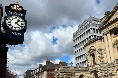 A black clock with a white face with the Norwich Union branding shows the time as 13:20, as dark clouds gather behind it.