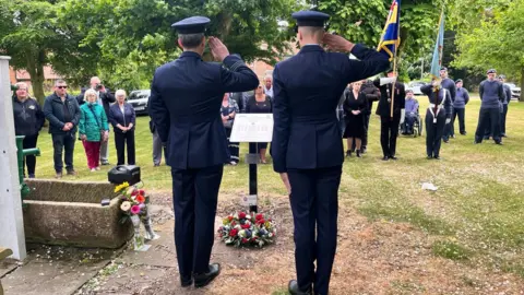 Two servicemen wearing blue uniforms salute in front of a white plaque mounted on a black stand. A wreath of white and red roses and other flowers have been laid at its base. A small crowds of people, including people dressed in uniforms with regimental colours, watch.
