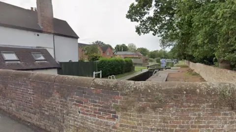 A view from a brick bridge which is built over a canal. The view shoes the canal stretching off into the distance with trees to the right of it and red brick houses to the left. 