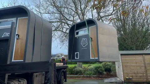 BBC Two grey pods being delivered - one is being craned from a lorry to the ground. Each has a small window and brown door with keypad and Amazing Grace Spaces logo on the front.