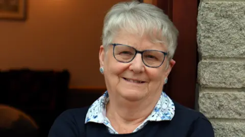 Charlie Gilmour/BBC A lady in her 70s smiles at the camera, wearing blue-framed glasses and two blue button earrings. She wears a white blouse with blue cornflowers on the collar and a navy jumper over the top.